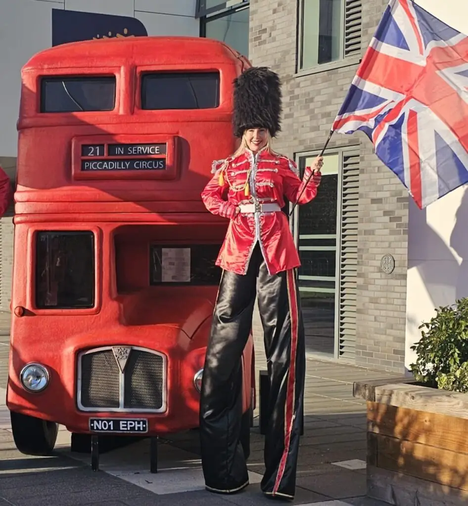 tall royal guard stilt walker in front of a red london bus waving a british flag